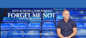 John Kenny sitting in a stadium of blue seats. Forget Me Not - For the Child You Left Behind, with John Kenny. Brick in the Wall Films.